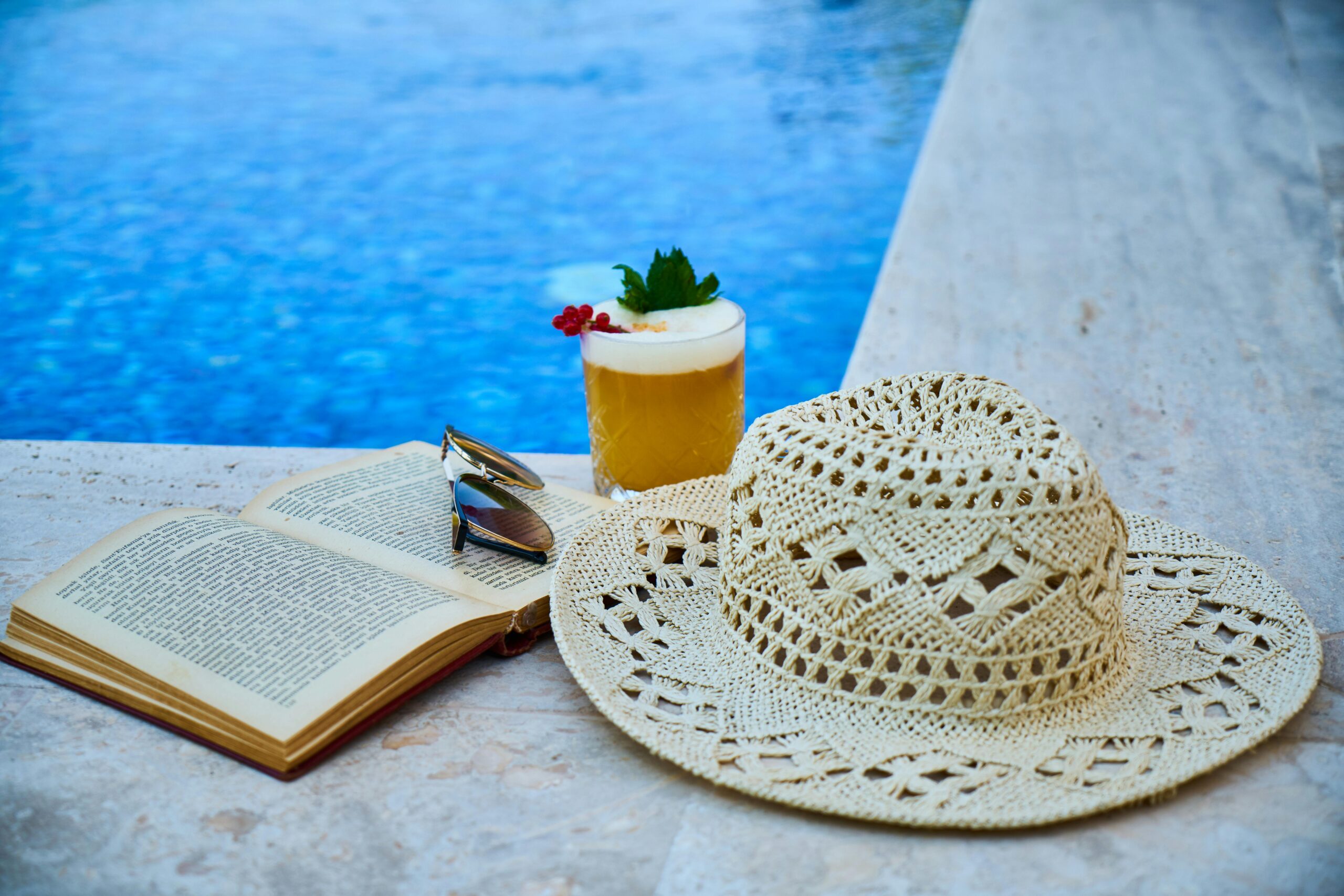 Enjoy a tranquil summer moment with a refreshing drink, hat, and book by the poolside.