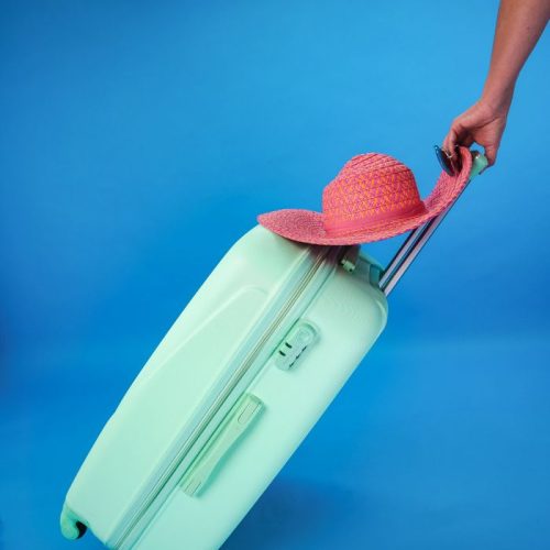 Mint green suitcase with pink sun hat against a blue background, evoking summer travel vibes.
